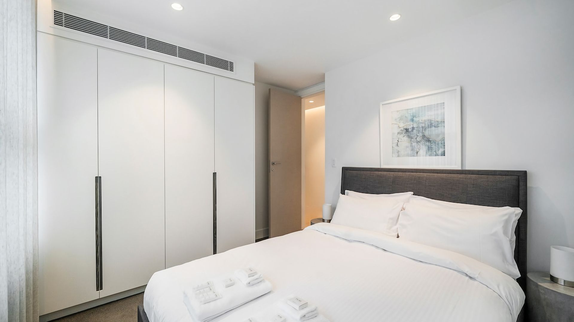 Ein Hotelzimmer mit einem Bett, einem Kleiderschrank, einem Nachttisch und einem gerahmten Kunstwerk an der Wand.