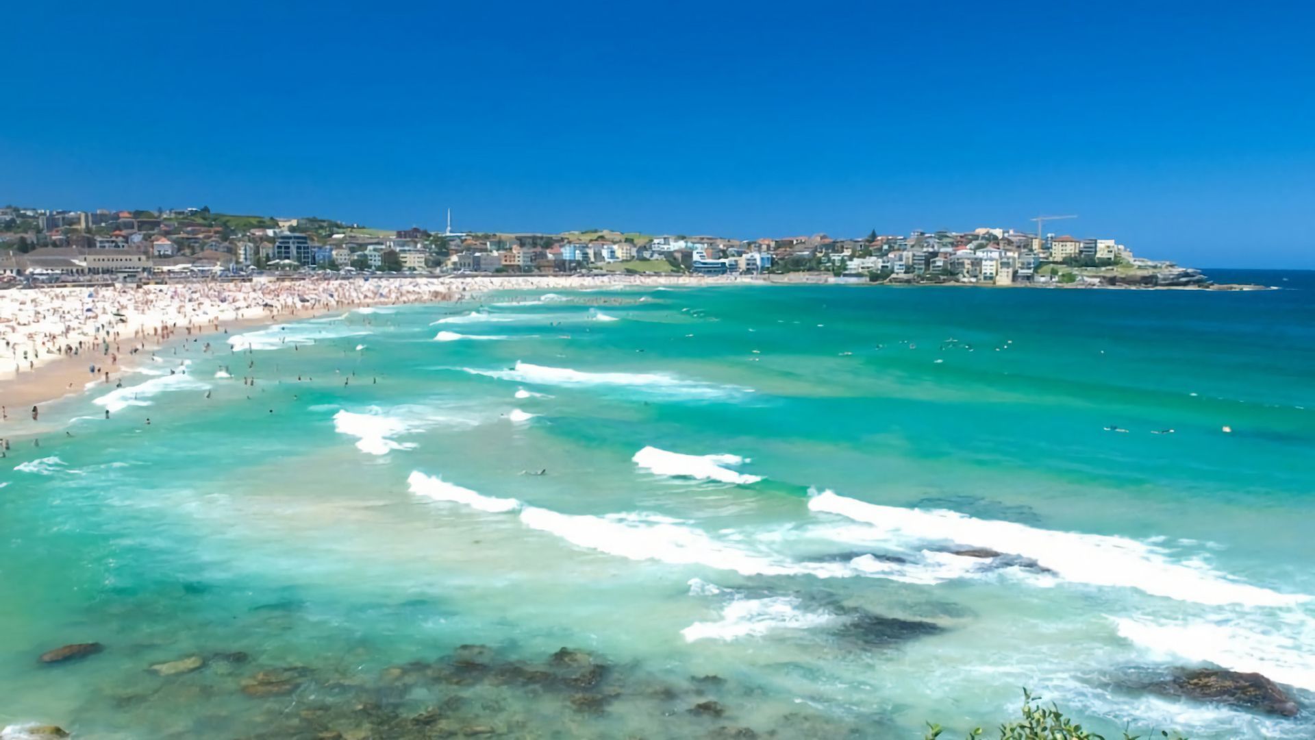 A coastal beach scene with clear turquoise waters, a sandy shore, and buildings in the background.