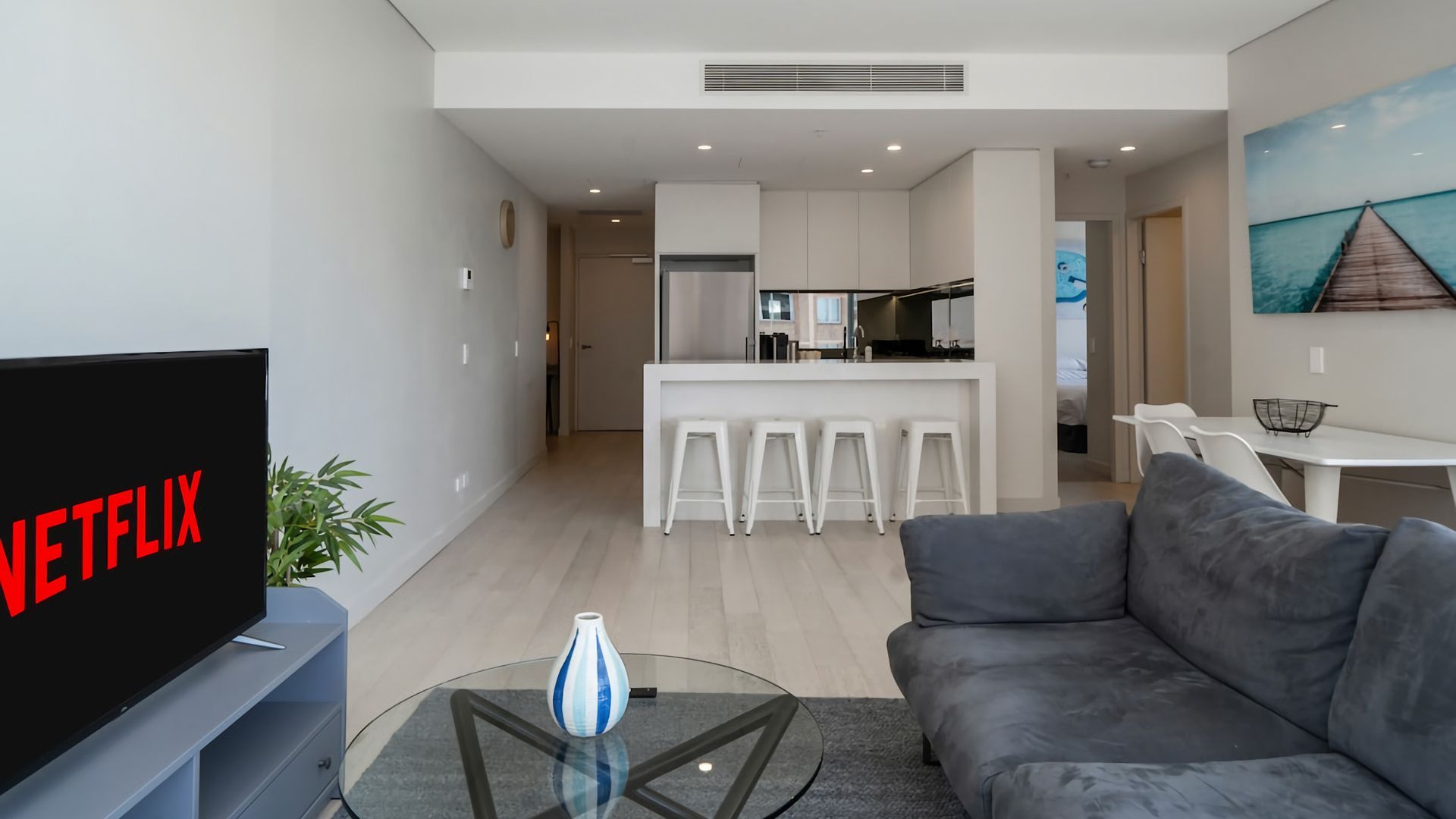 A modern living room with a flat-screen TV, a glass coffee table, and a grey sofa.
