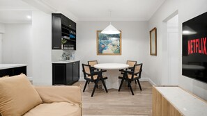 En moderne stue med et rundt spisebord, sorte stole, en beige sofa, et sort skab med glas og et maleri på væggen, samt et Netflix skilt.