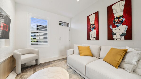 A modern living room with a white sofa, a small armchair, and a coffee table. There are two paintings on the wall and a window with blinds. - Luxury Paddington Terrace with Courtyard & Balcony (Paddington)
