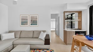 En moderne stue med en beige sofa, et spisebord med stole og et køkkenområde med et glaskab.