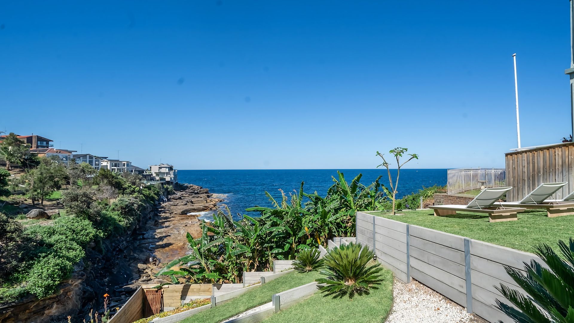 A coastal property with a view of the sea, a wooden deck with lounge chairs, and a rocky shoreline.