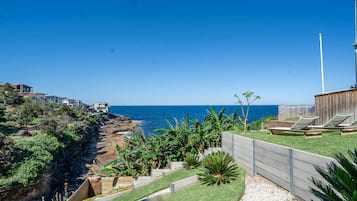 A coastal property with a view of the sea, a wooden deck with lounge chairs, and a rocky shoreline.