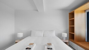 A modern hotel room with a large bed, two bedside tables, a wooden shelf unit, and a view of the outside.