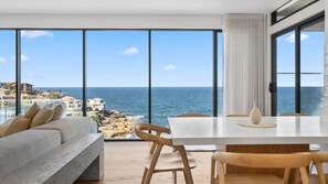 A modern living room with a large window overlooking the sea, a white sofa, wooden chairs, and a dining table. - South Coogee Escape, Heated Pool & Spacious Living (Maroubra)