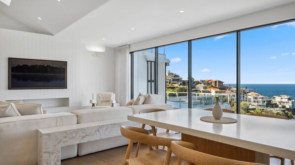A modern living room with a large window, a white sofa, and a dining area with wooden chairs.