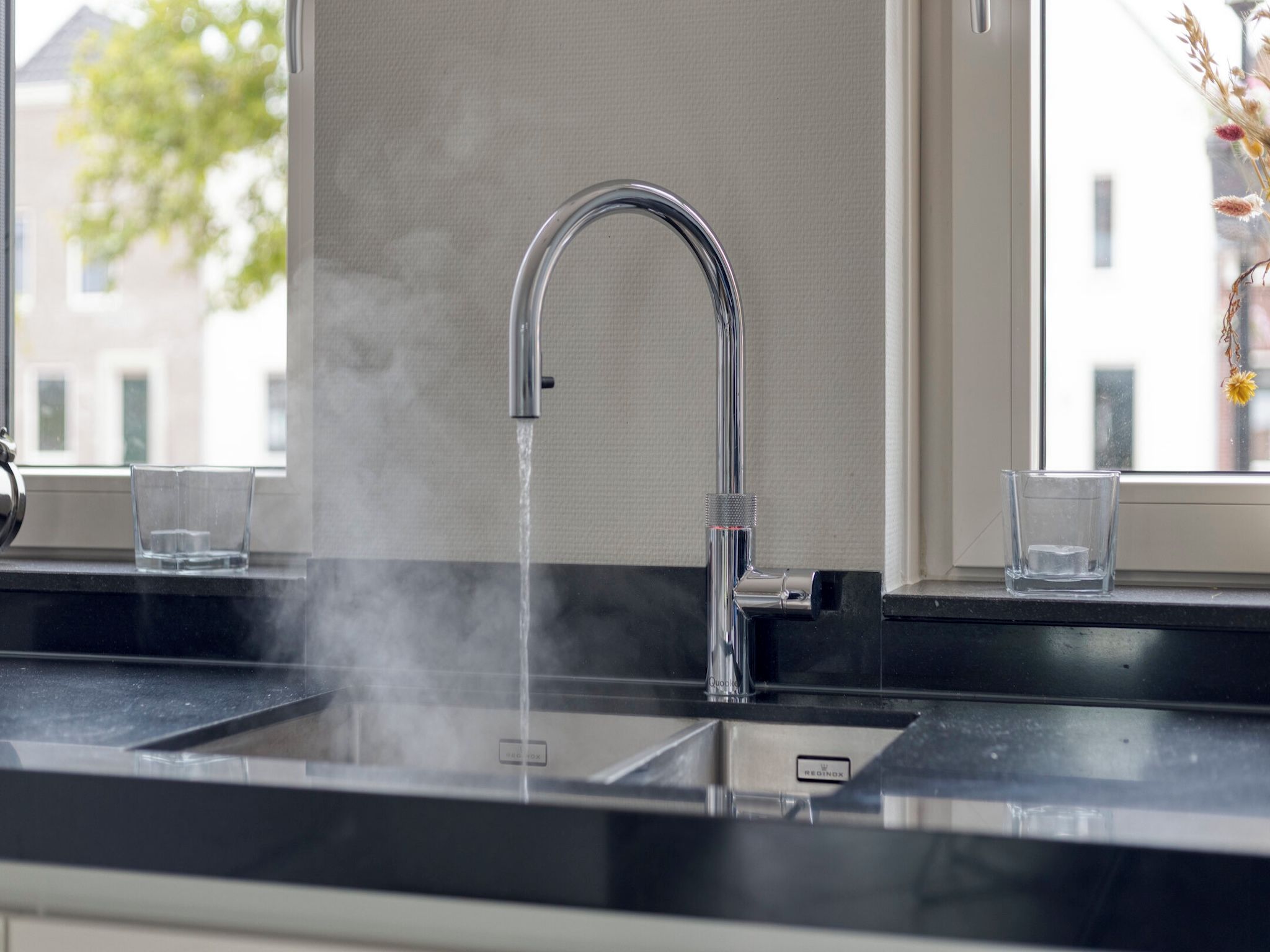 Een moderne keuken met een roestvrijstalen kraan, twee glazen op het aanrecht en een raam met uitzicht op gebouwen en bomen.