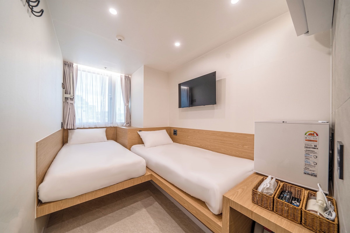 A hotel room with two single beds, a flat-screen TV mounted on the wall, a refrigerator, and a window with curtains.