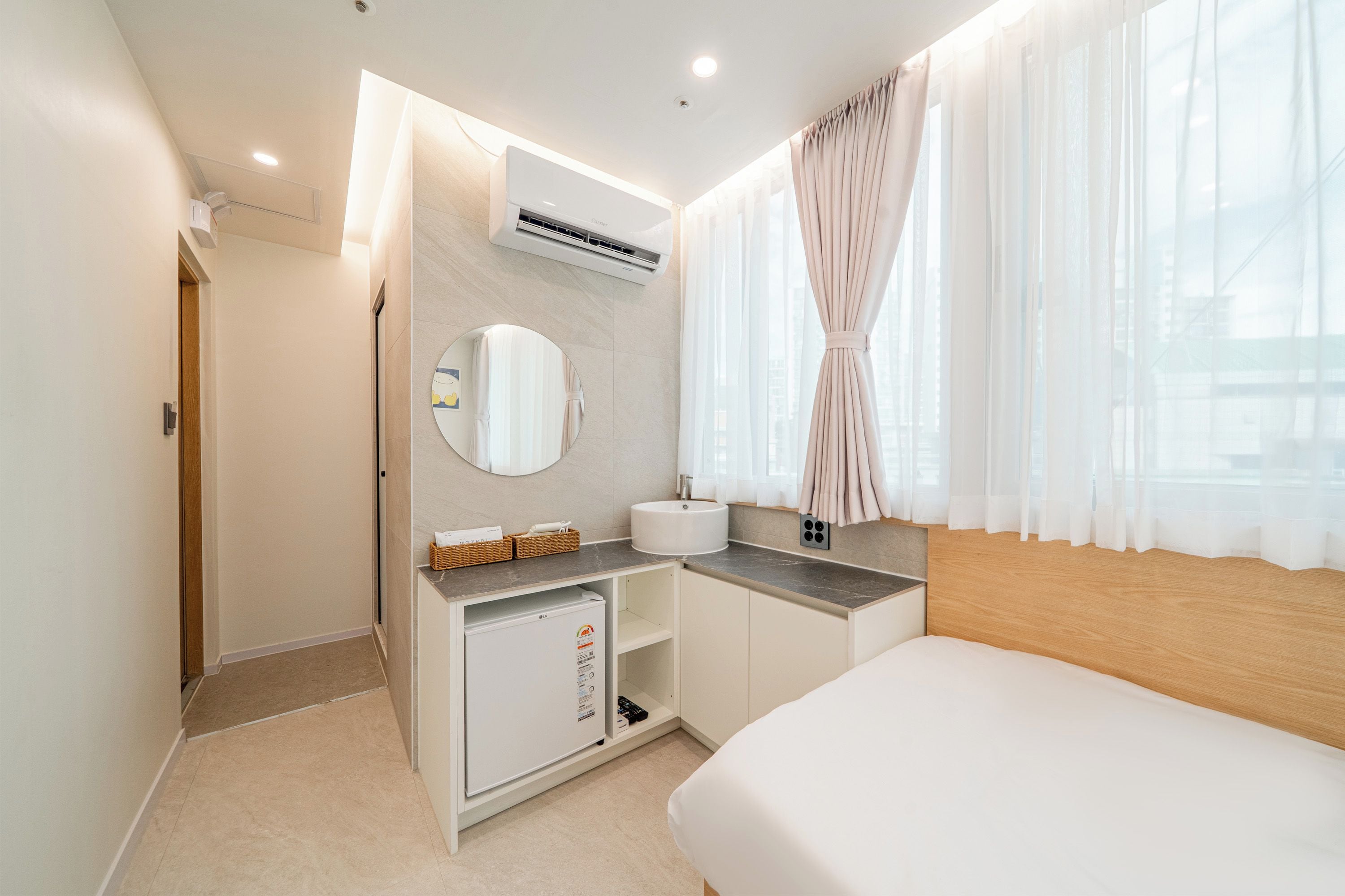 A modern hotel room with a bed, a small kitchenette, a bathroom, and a large window with sheer curtains.