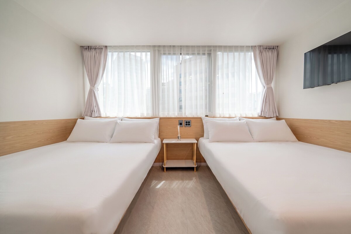 A hotel room with two beds, a wooden headboard, a small bedside table, and a window with sheer curtains.