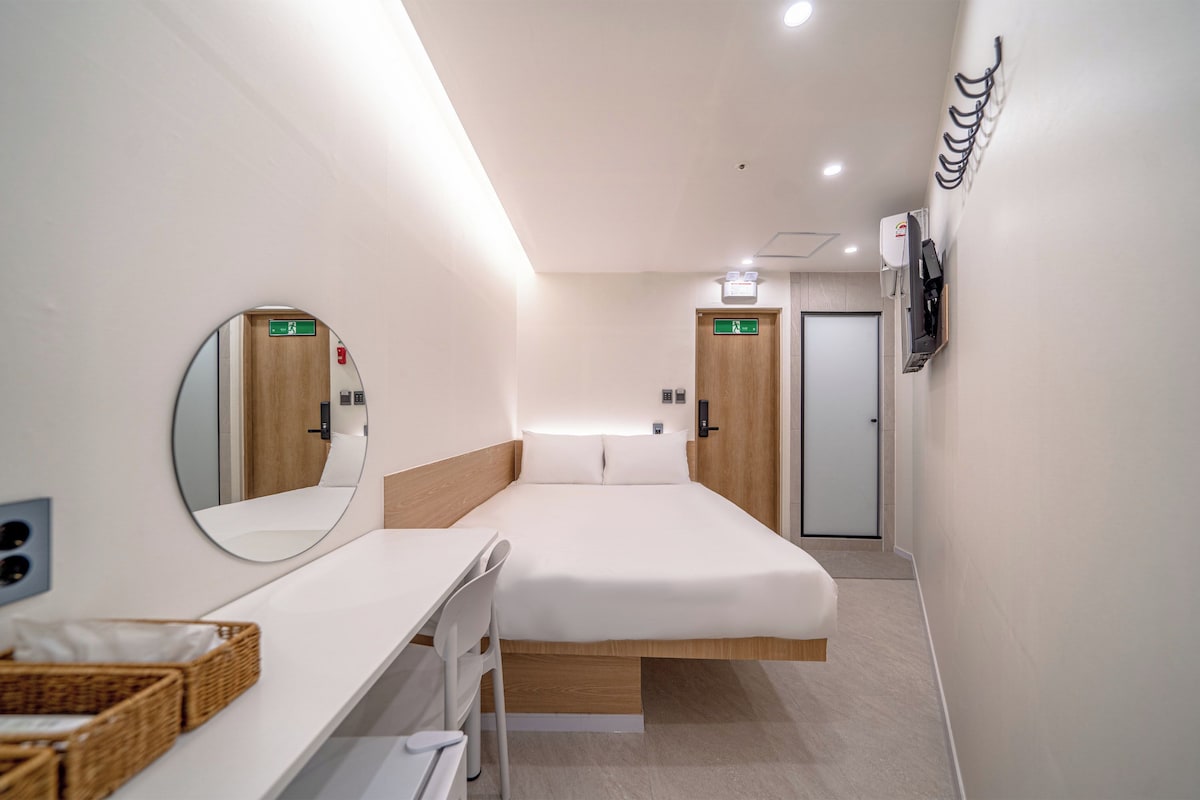 A modern hotel room with a bed, a desk, a mirror, a wardrobe, and a door with an emergency exit sign.
