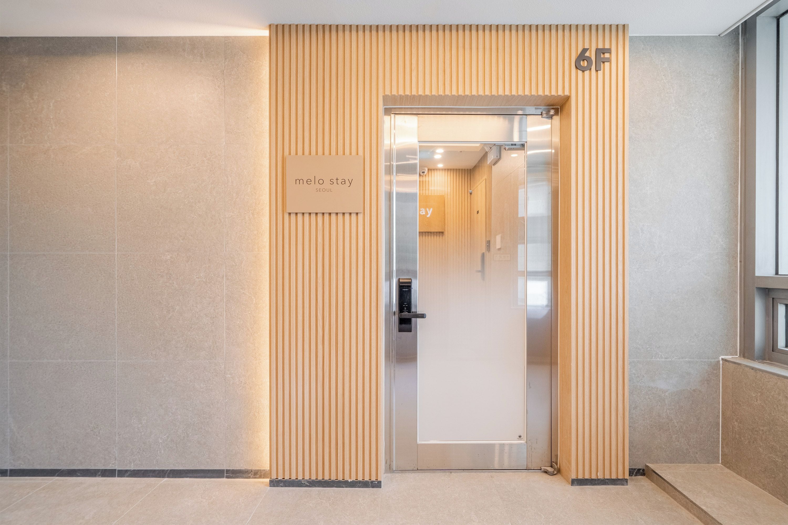 Une entrée d’hôtel moderne avec un décor de panneaux en bois, une enseigne indiquant « melo stay Séoul » et une porte portant le numéro « 6F ».