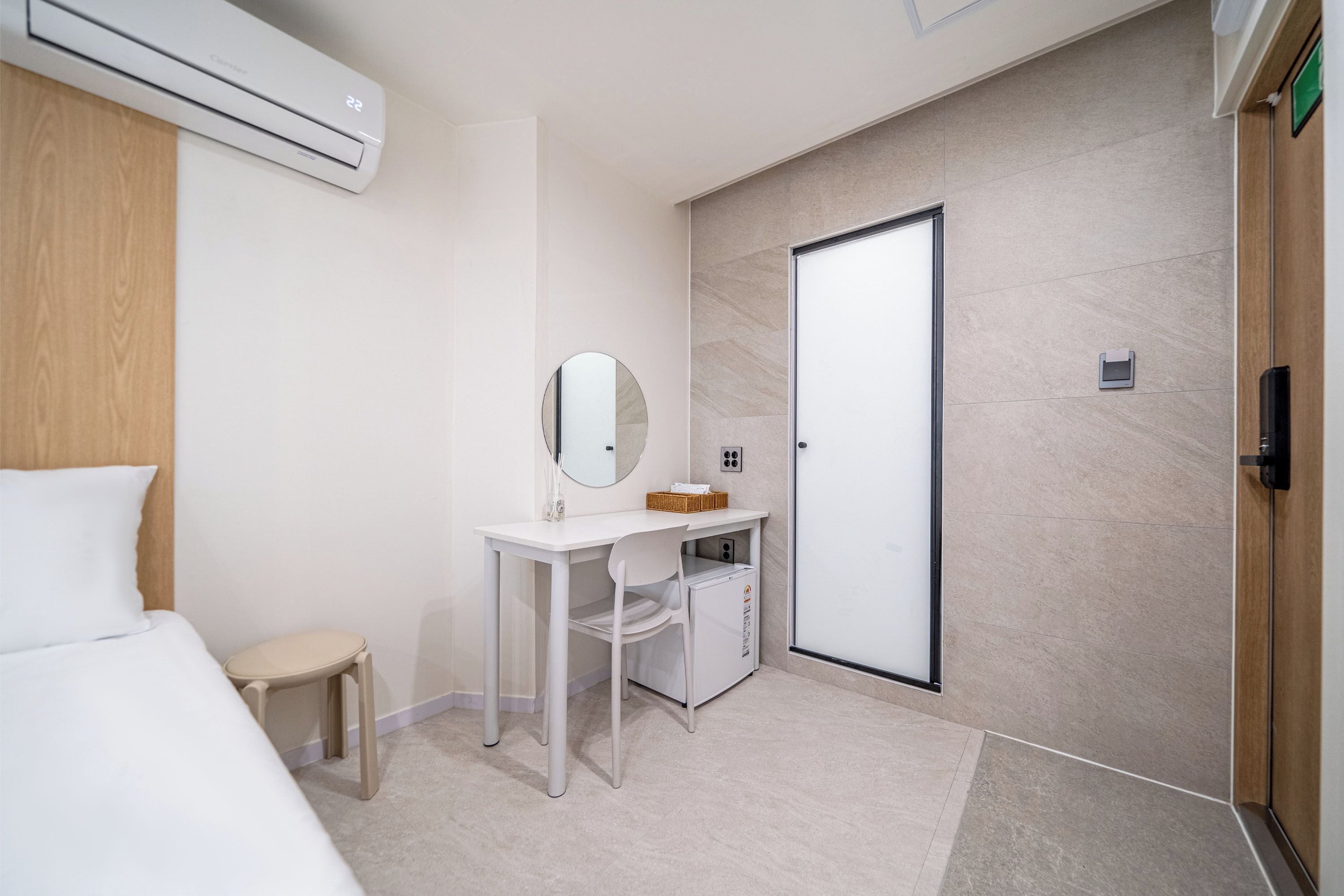 A modern hotel room with a bed, a desk, a mirror, a small cabinet, and a door.
