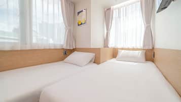 A hotel room with two single beds, a wooden headboard, a wall-mounted air conditioning unit, and sheer curtains.