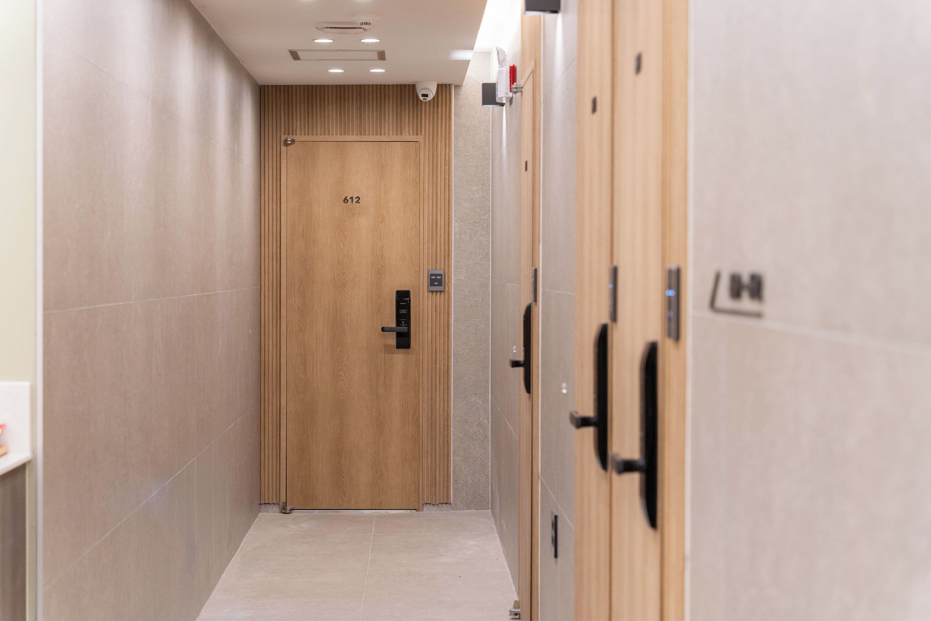 A hallway with a wooden door marked with the number 612, a security camera above, and a fire alarm on the wall.