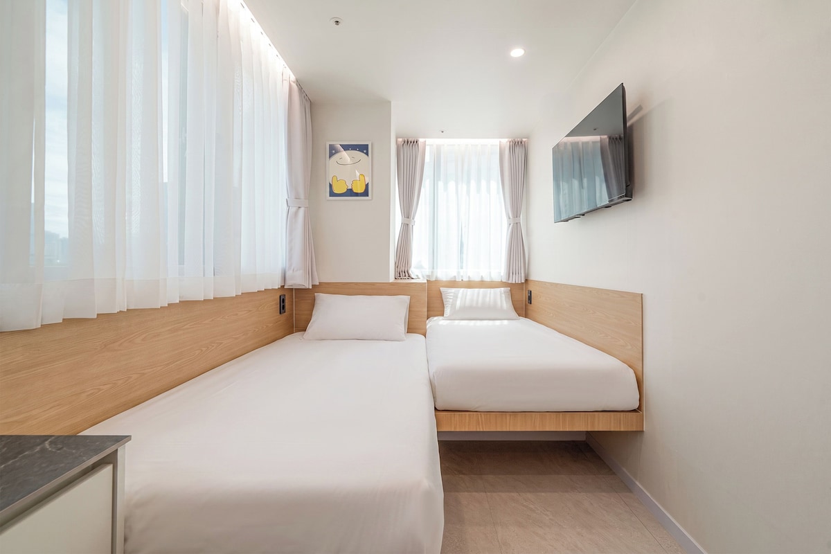 A hotel room with two single beds, a TV mounted on the wall, and a window with sheer curtains.