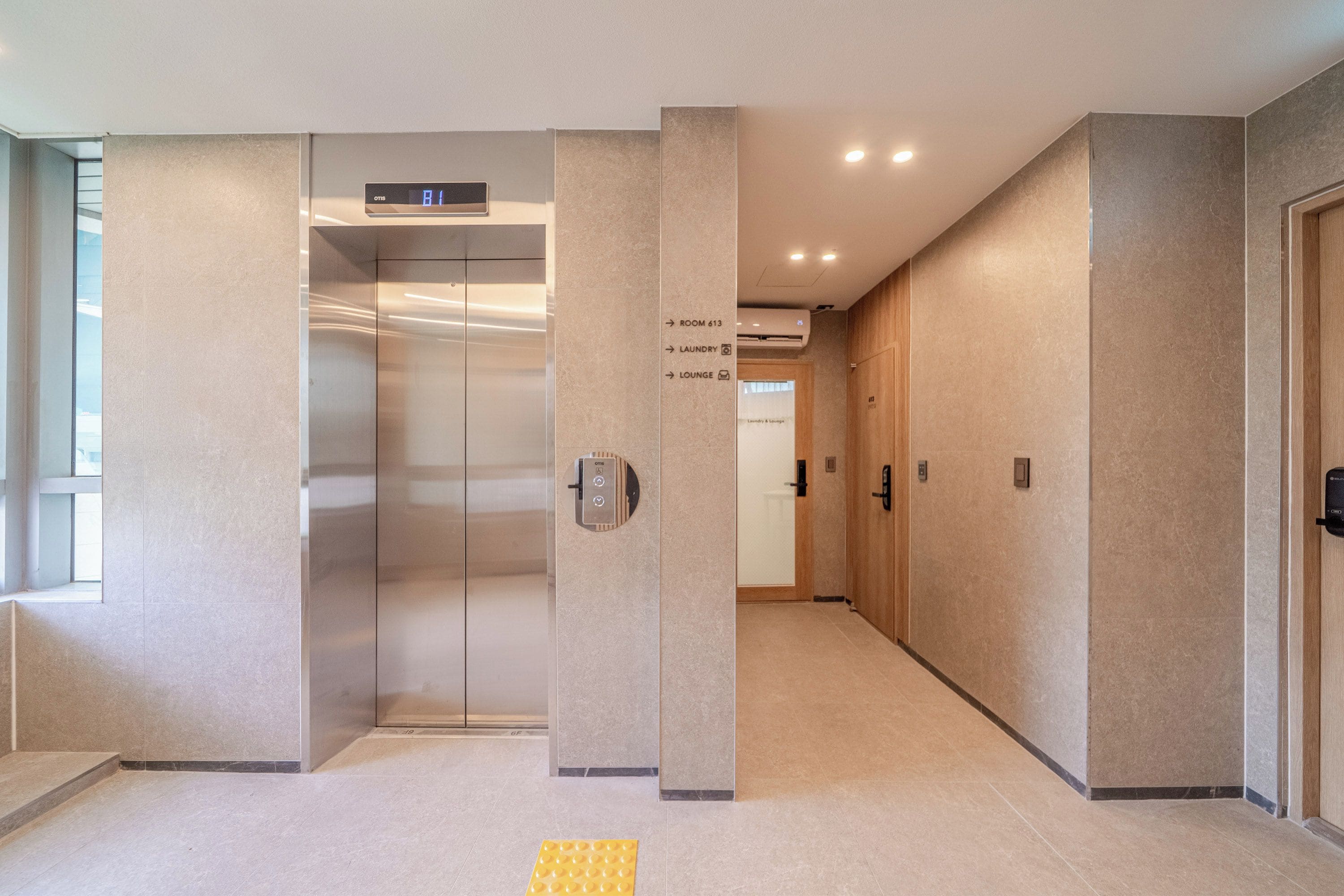Um elevador moderno com tela digital, sinalização clara indicando o caminho para o lounge e faixa tátil no chão.