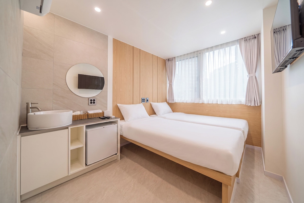 A modern hotel room with a large bed, a bedside table, a built-in TV, and a bathroom with a sink and mirror.