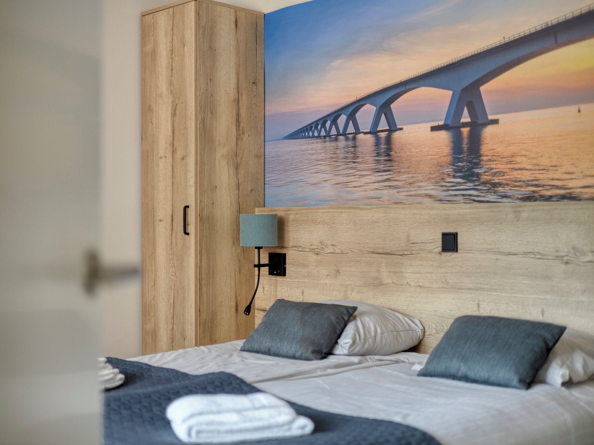 A bedroom with a bed, pillows, a nightstand, a wooden wardrobe, and a wall-mounted lamp.