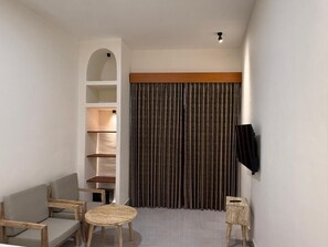 A hotel room with a sofa, a small round table, a TV mounted on the wall, and a built-in shelving unit. - Villa Basheera (Gili Trawangan)
