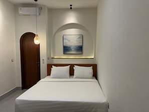 Ein Hotelzimmer mit einem Bett, einer Holztür, einer Klimaanlage, einer Pendelleuchte und einem gerahmten Bild an der Wand.