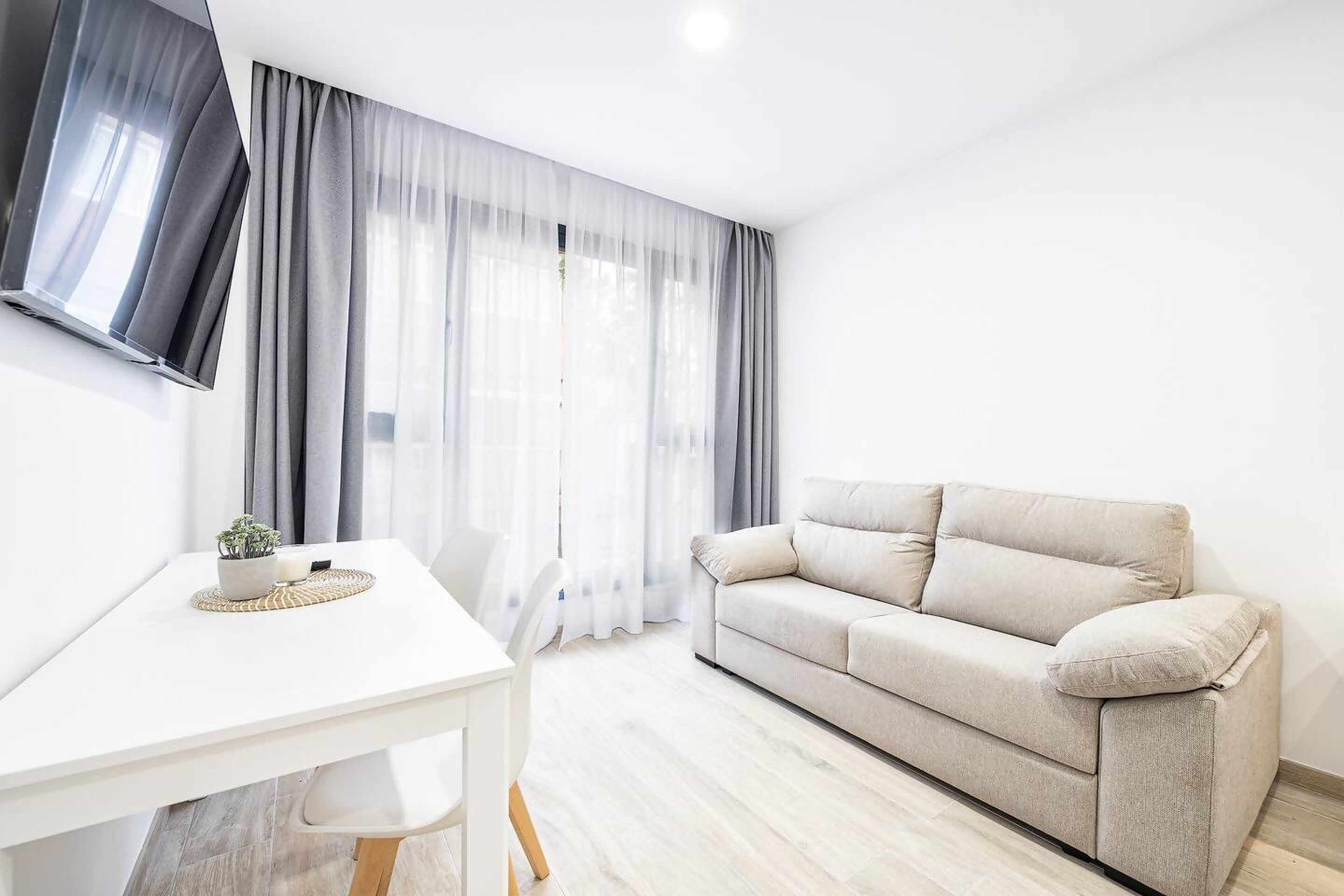 A modern living room with a white sofa, a dining table, and a TV mounted on the wall.