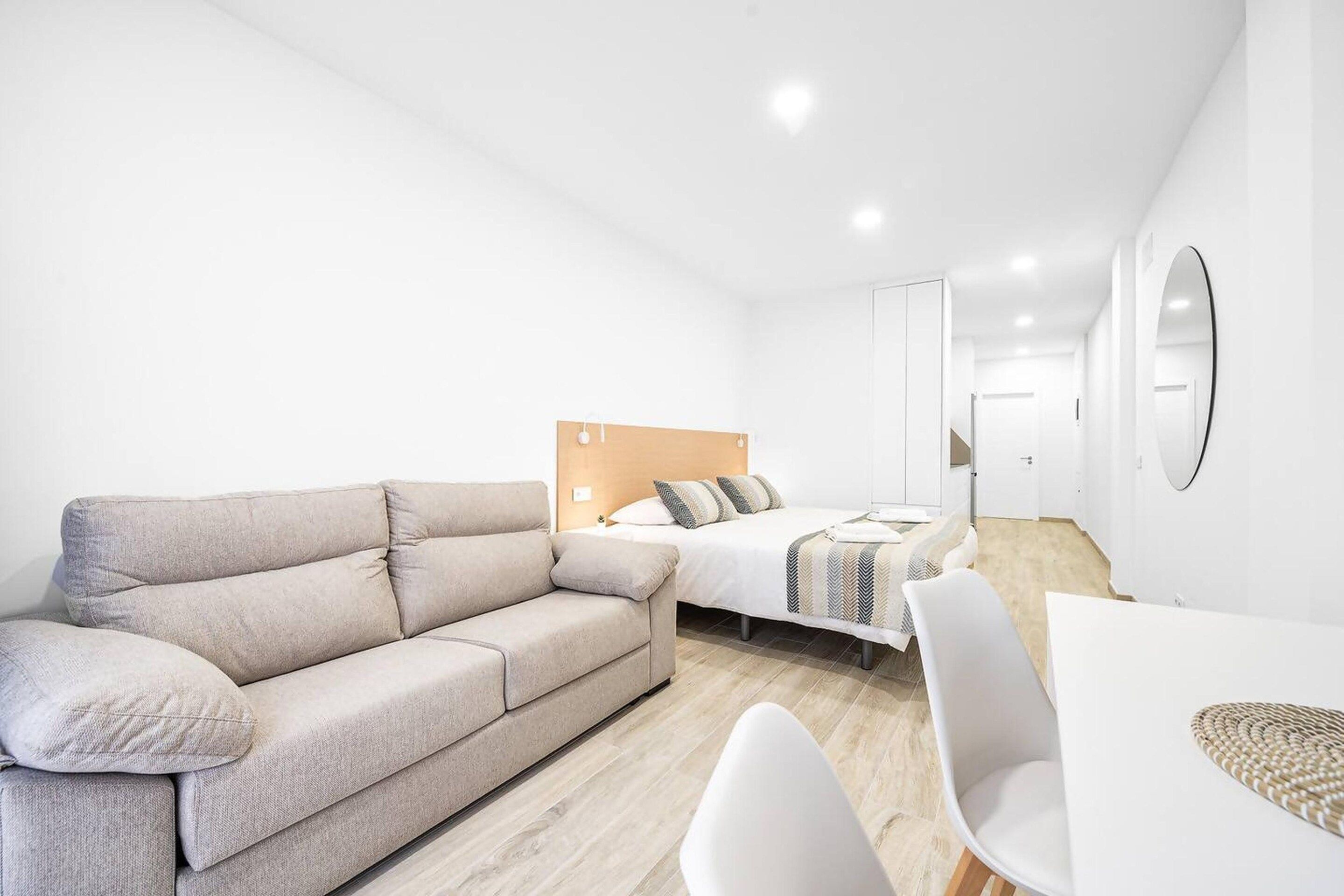 A modern, minimalist hotel room with a sofa, a bed with a wooden headboard, a dining table, and a large mirror.