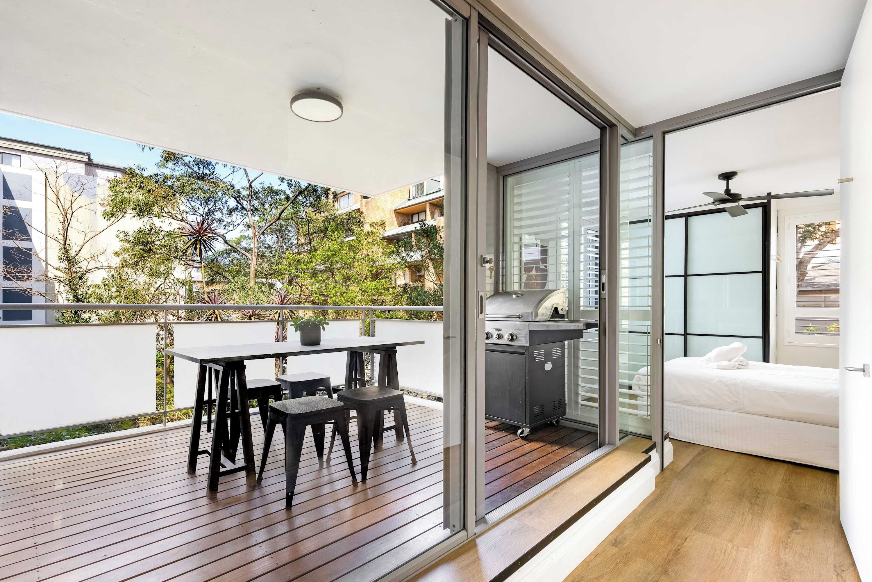A modern balcony with a wooden deck, a dining table with chairs, a grill, and a white sofa.