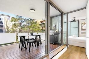A modern balcony with a wooden deck, a dining table with chairs, a grill, and a white sofa. - The Albion Courtyard, Surry Hills (Surry Hills)