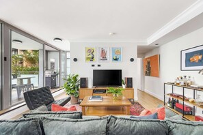 A modern living room with a flat-screen TV, a wooden coffee table, and a sofa. There are framed posters on the wall, a bookshelf with various items, and a sliding glass door leading to an outdoor area with a barbecue. - The Albion Courtyard, Surry Hills (Surry Hills)