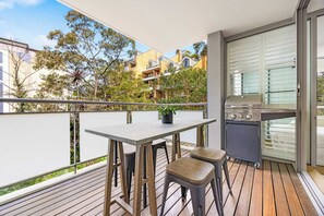 Un balcon avec une table et deux tabourets, un petit coin cuisine équipé d’un barbecue, et une vue sur les bâtiments et les arbres environnants.