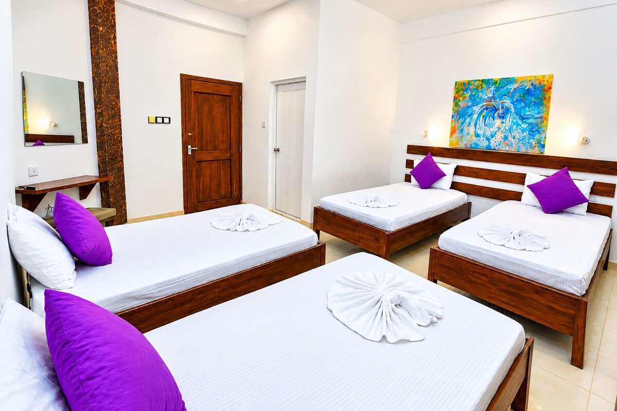 Habitación de hotel con tres camas, cada una con sábanas blancas y almohadas moradas. Se observa un cabecero de madera, un cuadro en la pared y una puerta de madera.
