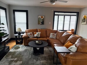 A living room with a brown leather sofa, a round coffee table, and a small side table. There are two windows with blinds, a ceiling fan, and artwork on the walls. - King Bed in Oak Park w/Chicago Chic (Oak Park)
