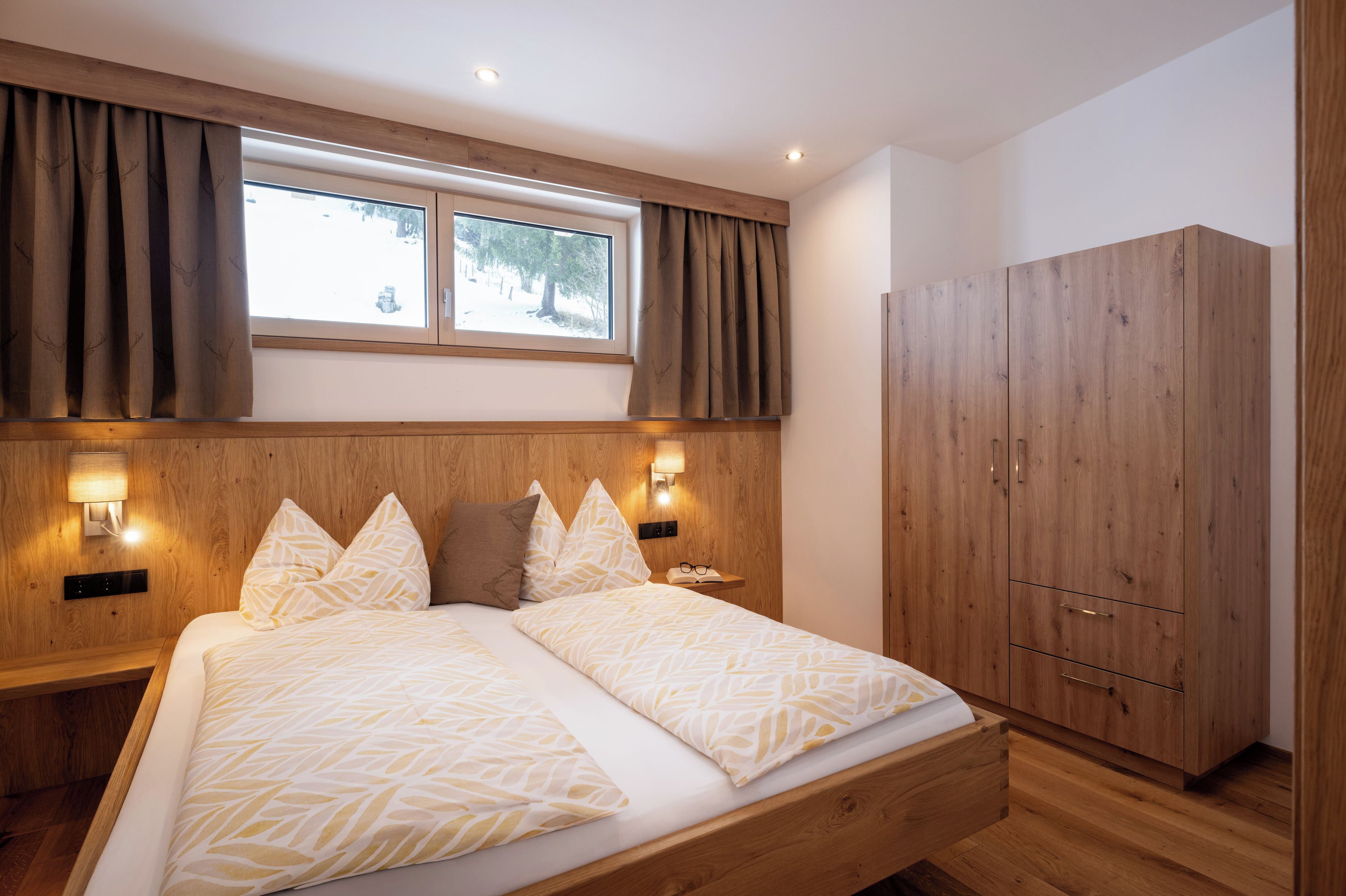 Une chambre à coucher avec un lit, une tête de lit en bois, deux lampes de chevet, une armoire et une fenêtre avec des rideaux.