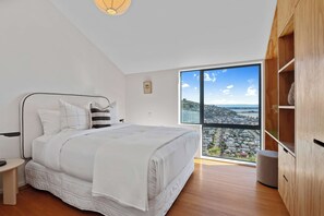 Ein Schlafzimmer mit einem großen Bett, einem Nachttisch, einem Holzschrank und Blick auf eine Küstenlandschaft durch ein großes Fenster.