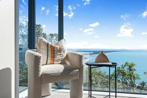A room with a view of a large body of water, a chair with a cushion, and a small table with a vase. - The Coastal Haven Villa (Christchurch)