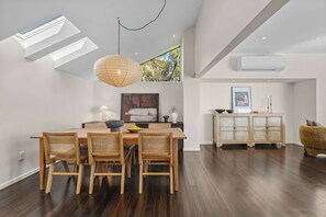 A modern dining area with a wooden table and chairs, a pendant light, and a large window with a view of trees. - The Coastal Haven Villa (Christchurch)