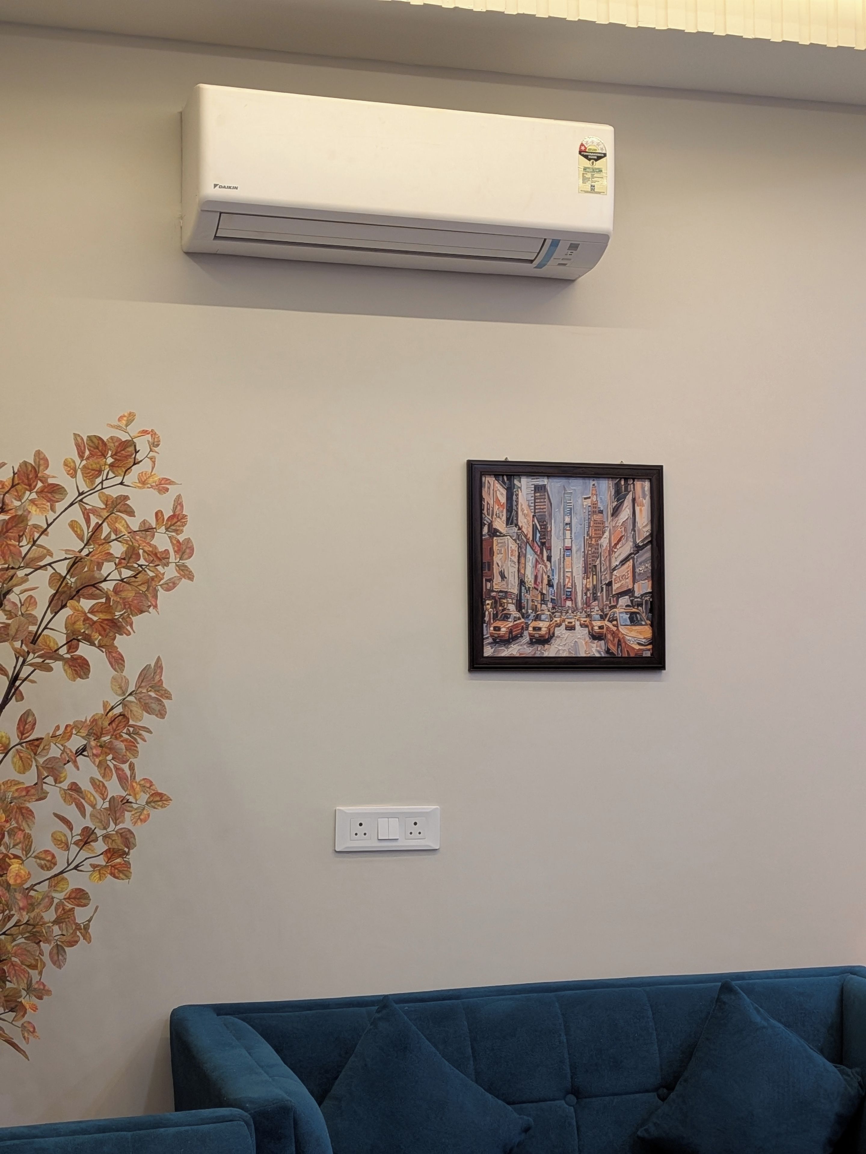 A room with a blue sofa, an air conditioning unit, a framed painting, and a wall-mounted electrical outlet.