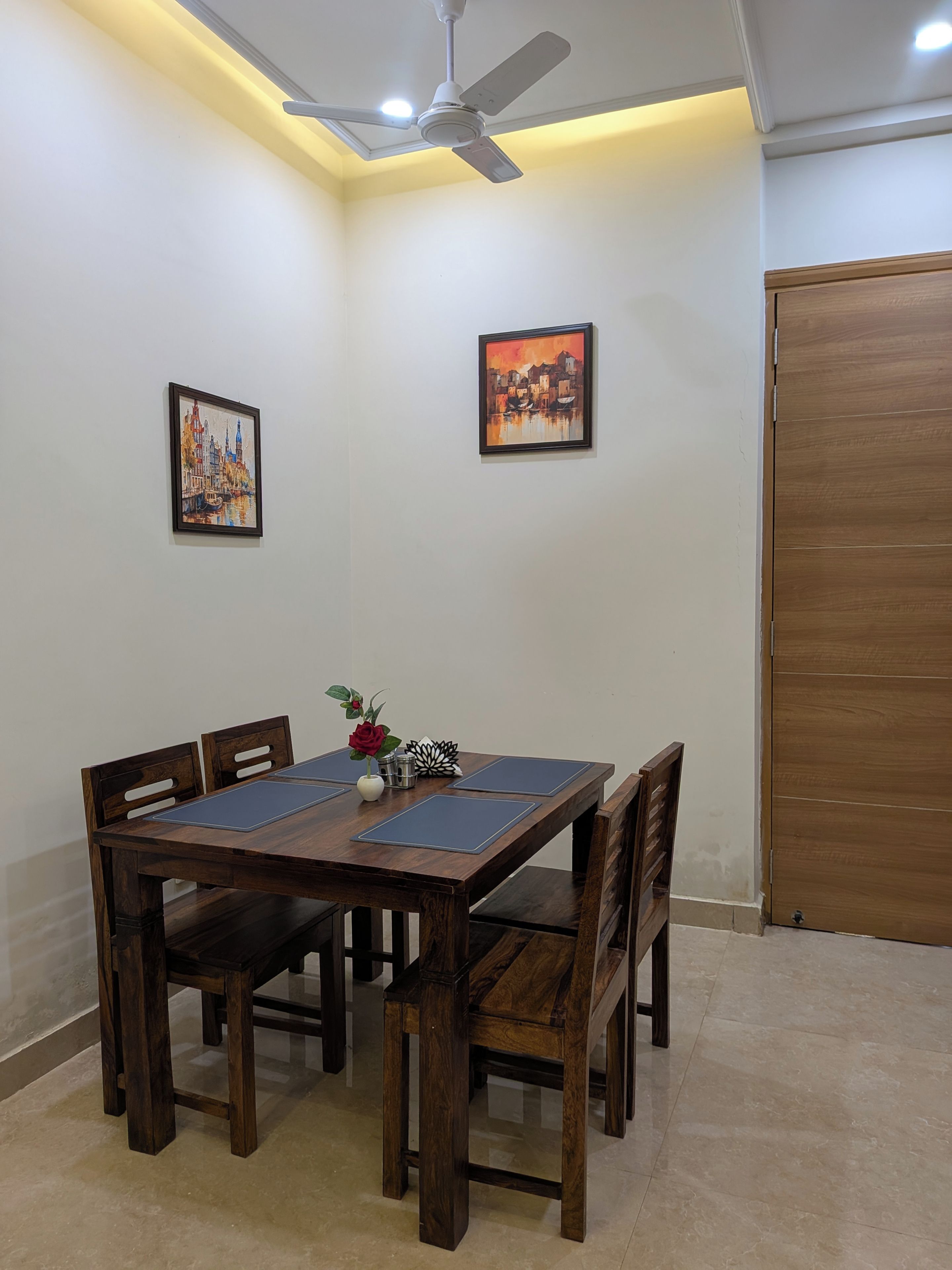 A dining area with a wooden table and chairs, two framed artworks on the wall, a ceiling fan, and a wooden door.