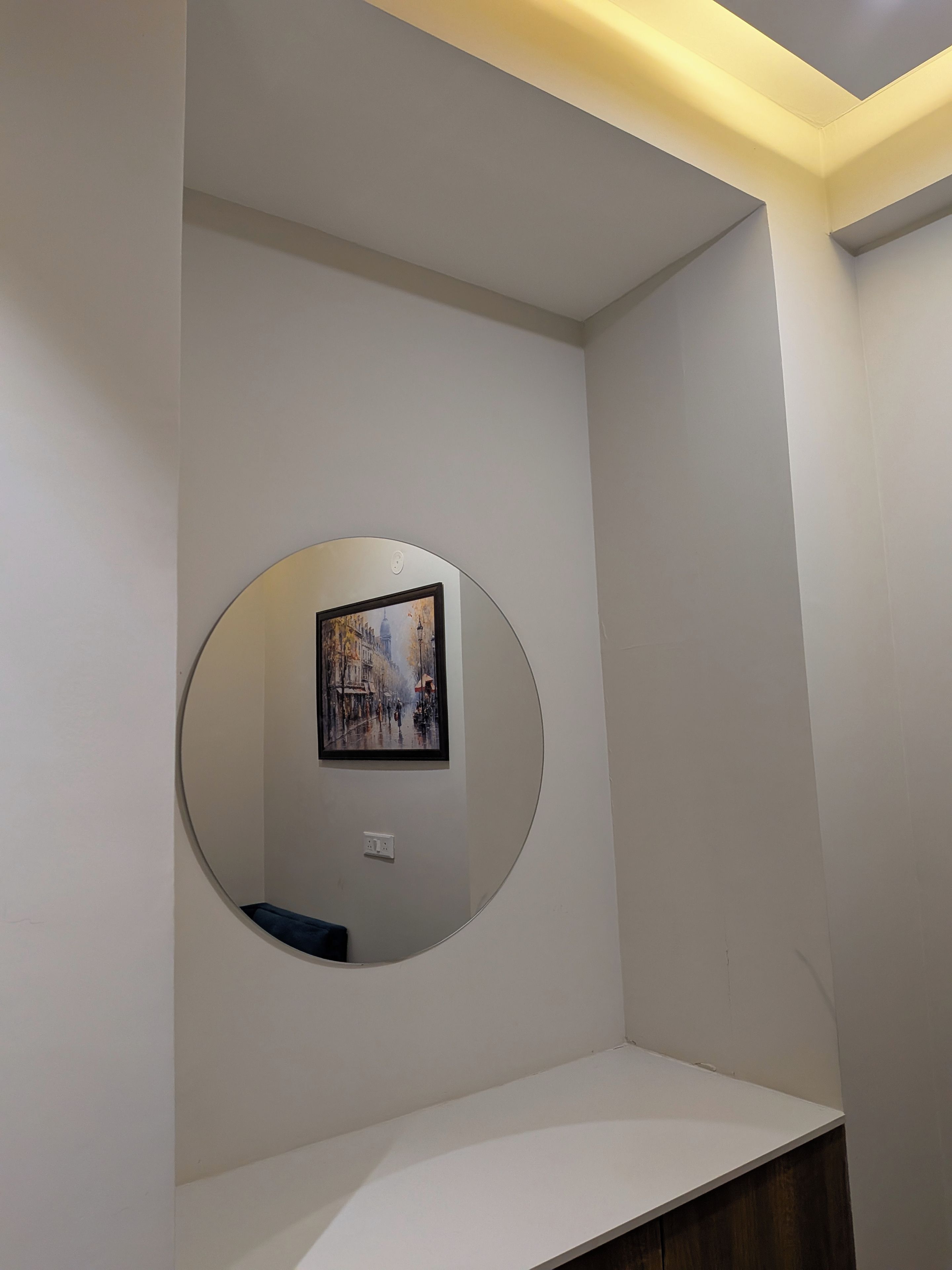 A room with a circular mirror on the wall, a white shelf, and a recessed ceiling light.