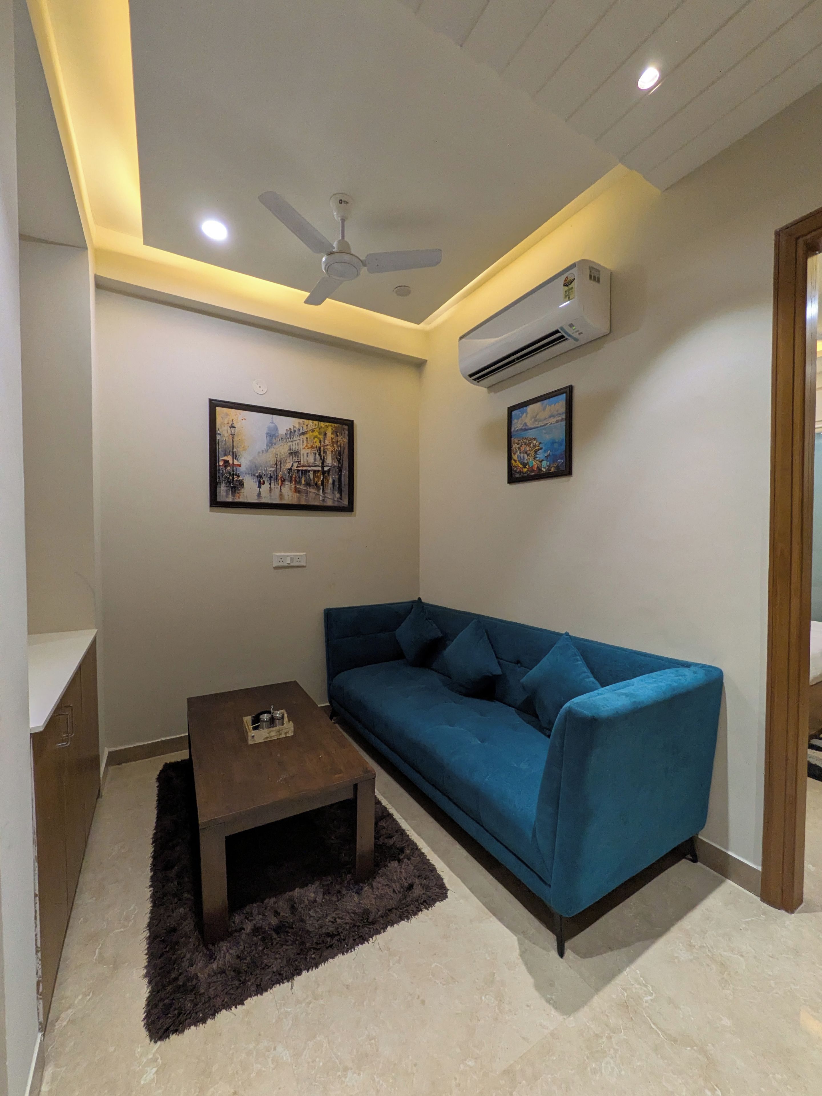 A living room with a blue sofa, a small table, a ceiling fan, and two wall-mounted air conditioners.