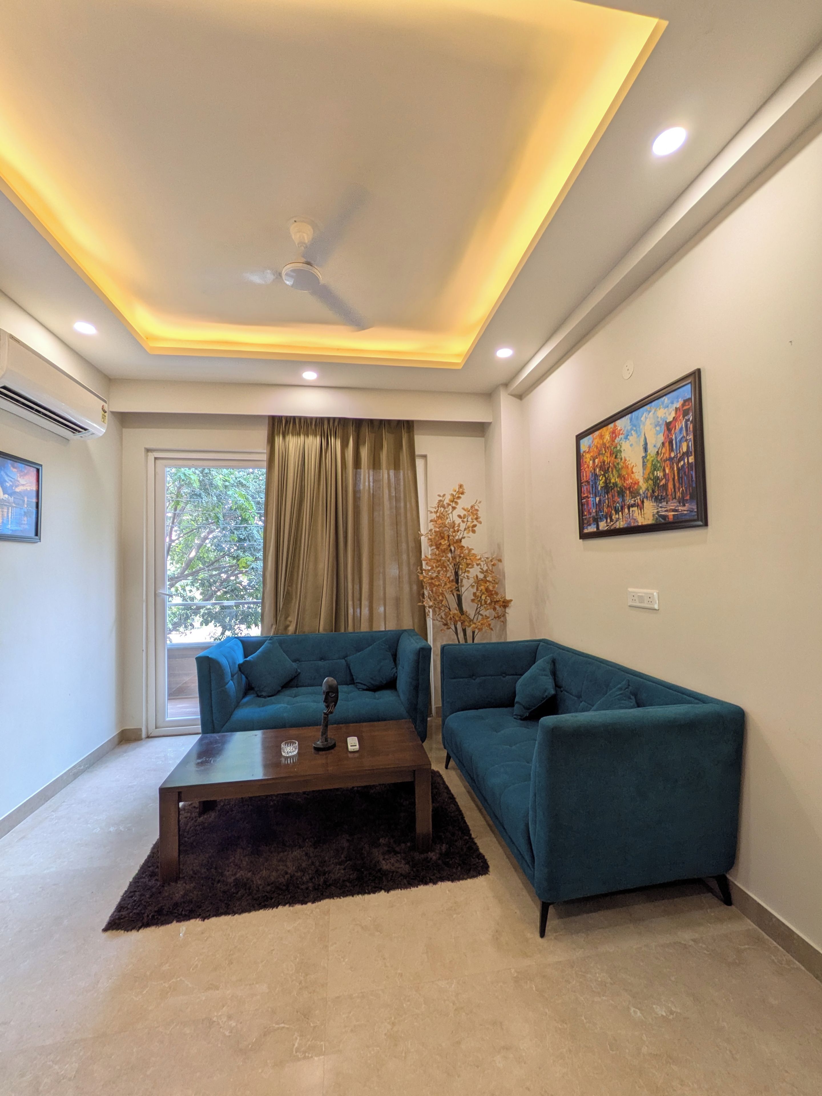 Un salon moderne avec un ventilateur de plafond, une grande fenêtre avec des rideaux, un canapé bleu, une table basse et un tableau accroché au mur.