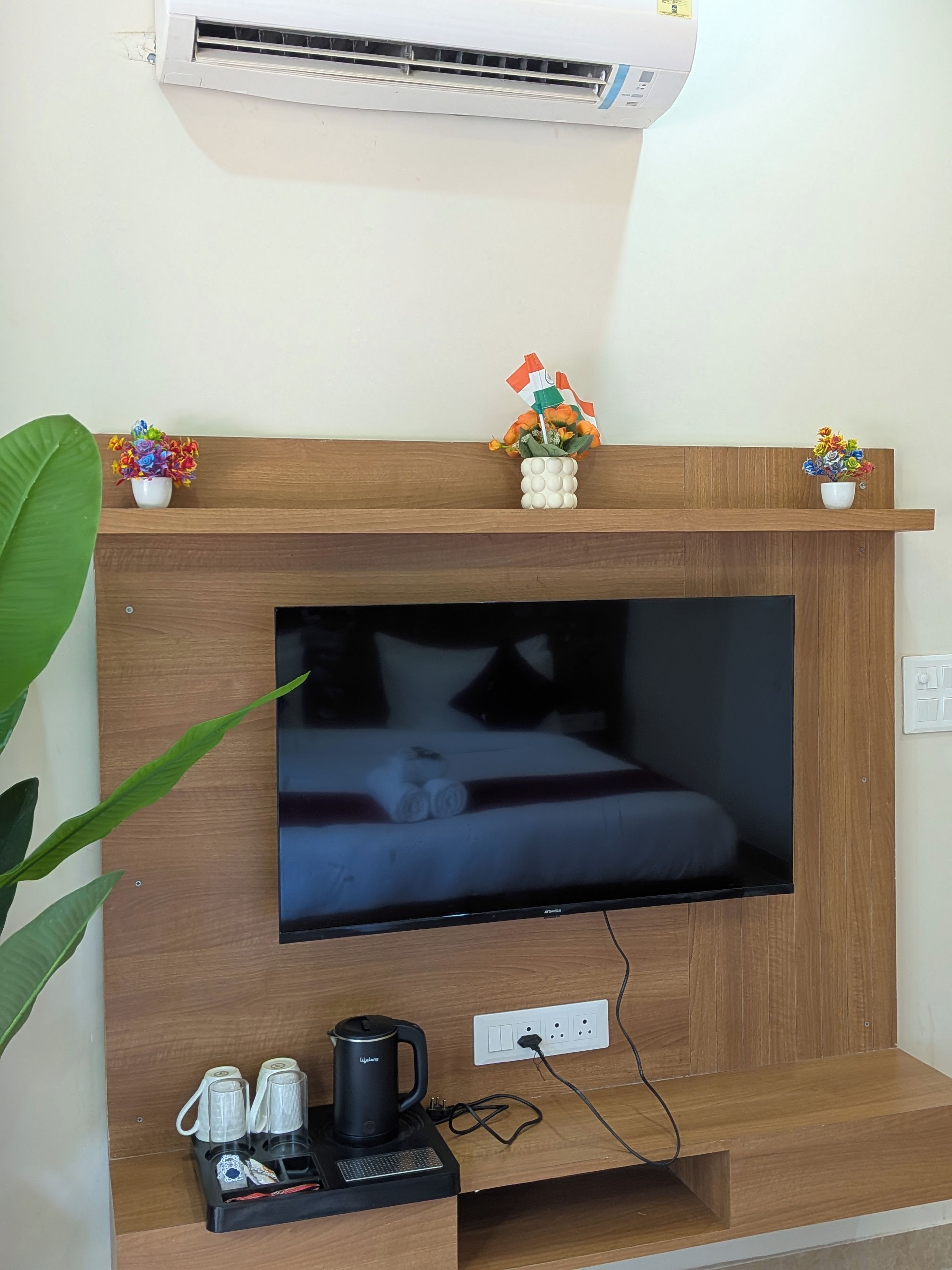 A room with a flat-screen TV mounted on a wooden wall, a small shelf with decorative items, a coffee maker, and a power outlet.