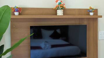 A room with a flat-screen TV mounted on a wooden wall, a small shelf with decorative items, a coffee maker, and a power outlet.