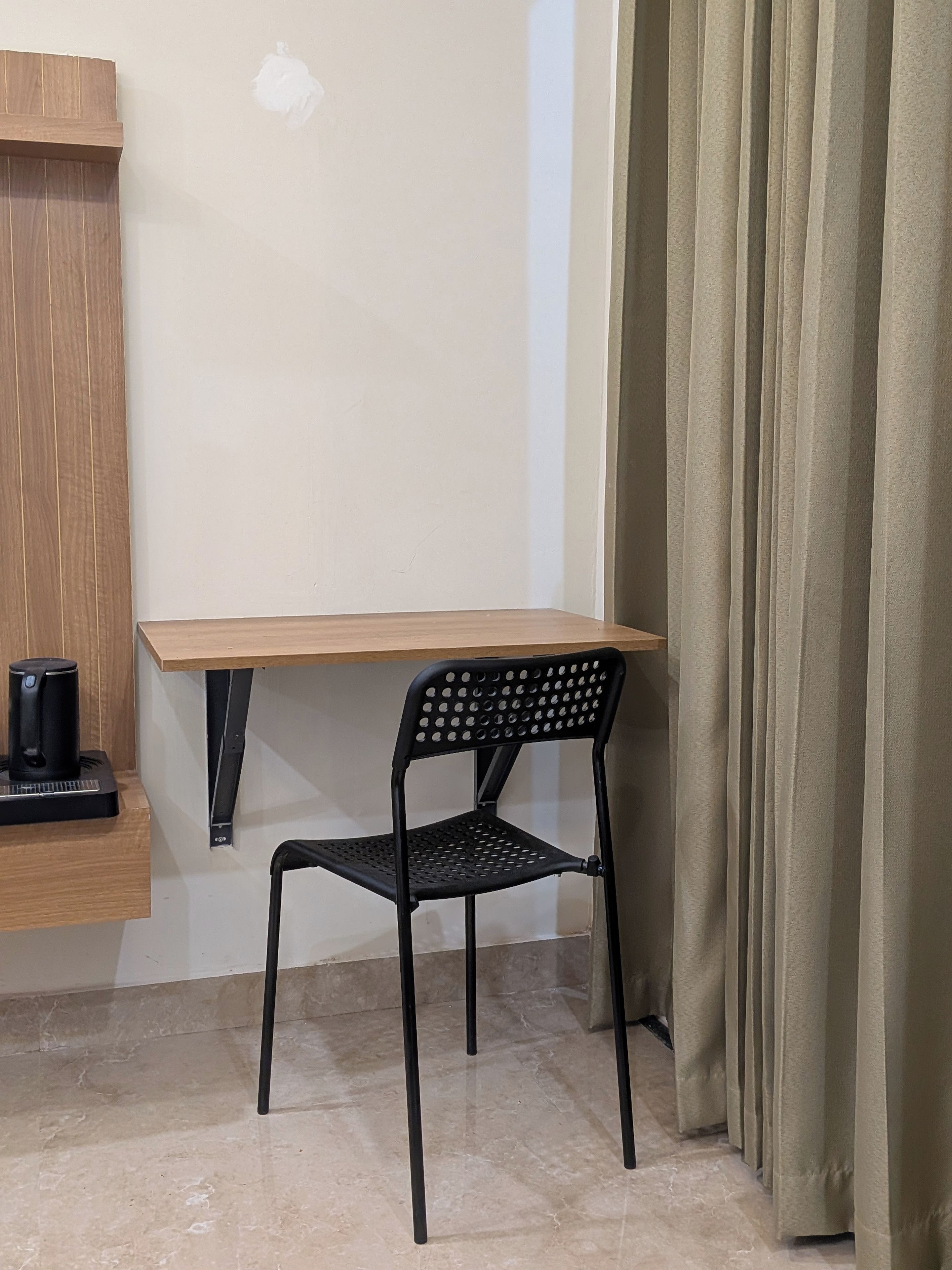 A small room with a wooden desk, a black metal chair, a coffee maker on the desk, and a curtain on the window.