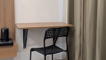 A small room with a wooden desk, a black metal chair, a coffee maker on the desk, and a curtain on the window.