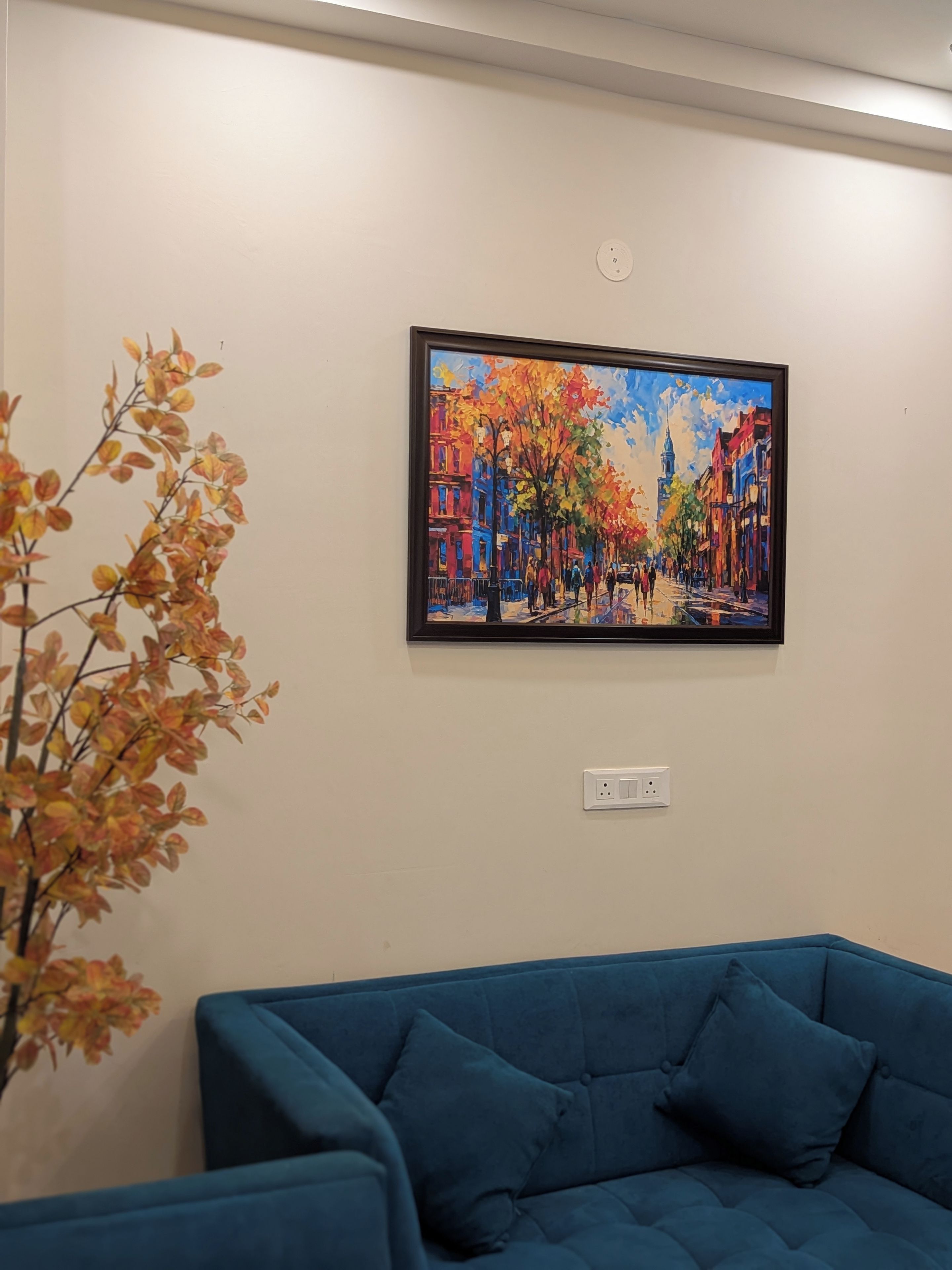 A room with a blue sofa, a painting of a colorful street scene, and a plant with autumn leaves.