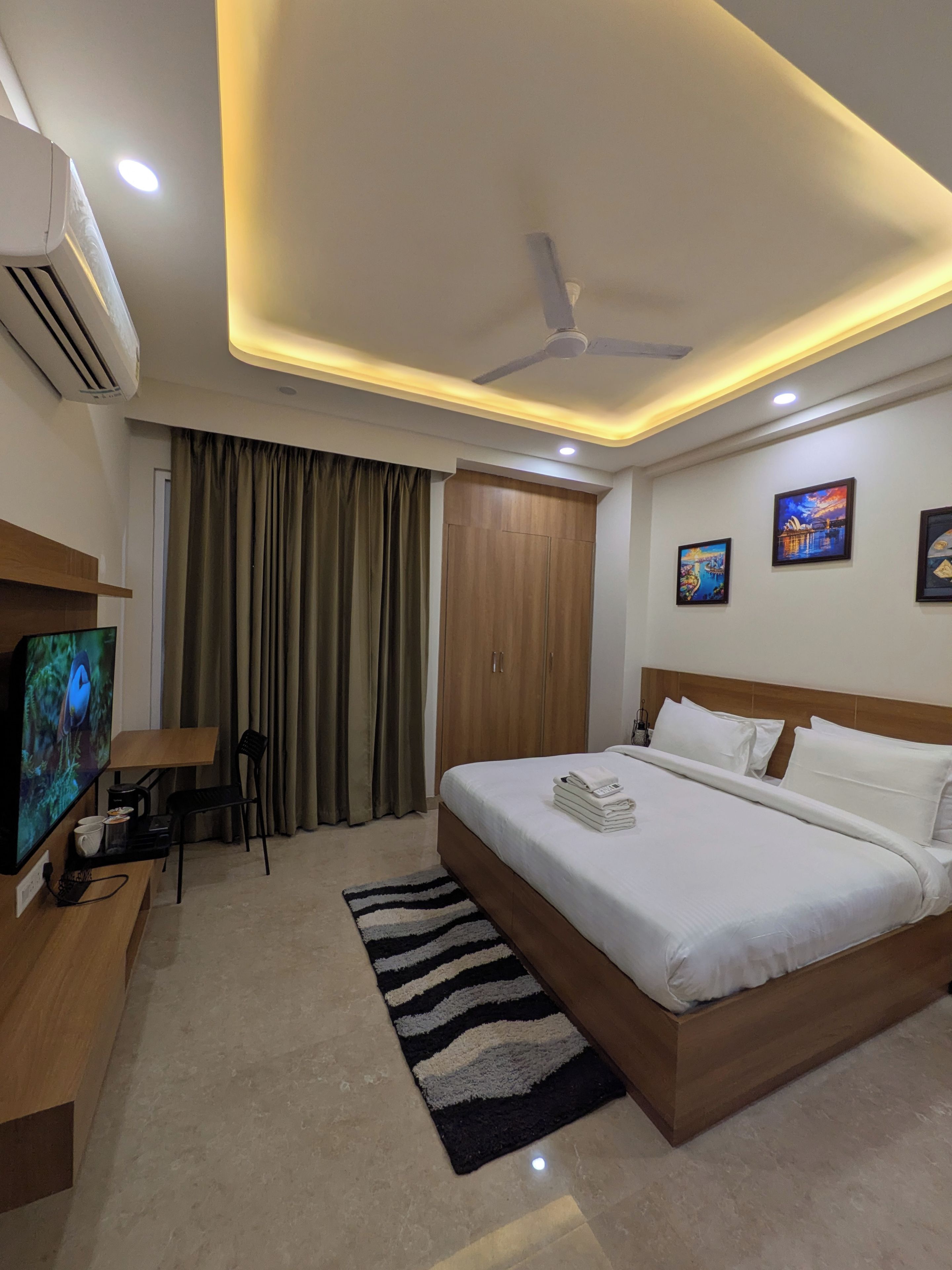 Quarto de hotel com cama, televisão, escrivaninha, cadeira, ventoinha no teto e porta.