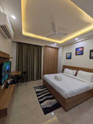 Quarto de hotel com cama, televisão, escrivaninha, cadeira, ventoinha no teto e porta.